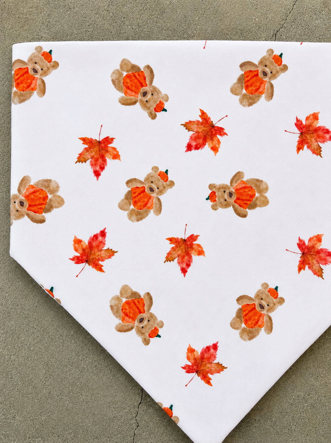 A square white bandana featuring a repeating pattern of watercolor autumn bears wearing orange pumpkins and red-orange maple leaves, laid flat on a textured gray surface.