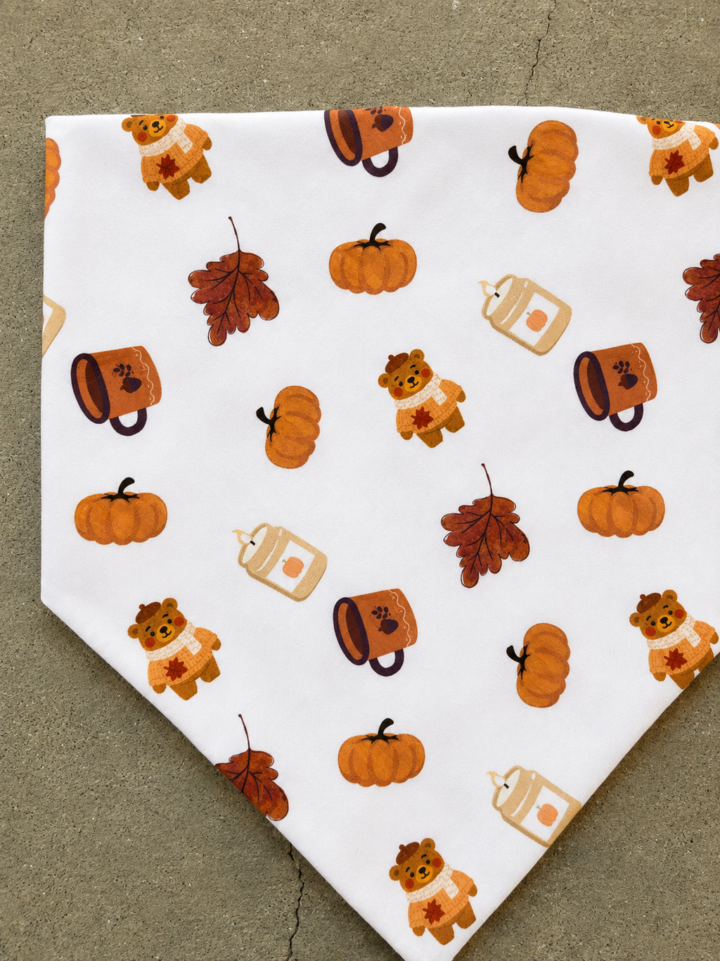A white bandana-style pet collar featuring a repeating autumn pattern of orange pumpkins, brown leaves, and teddy bears wearing orange sweaters, laid flat on a gray concrete surface.
