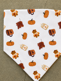 A white bandana-style pet collar featuring a repeating autumn pattern of orange pumpkins, brown leaves, and teddy bears wearing orange sweaters, laid flat on a gray concrete surface.