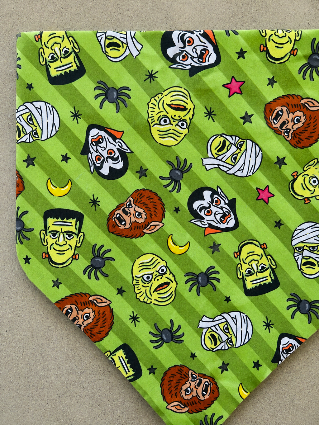 A green pet bandana featuring a Halloween-themed print with cartoon monster faces, black spiders, black stars, and yellow crescent moons on a diagonal striped background.