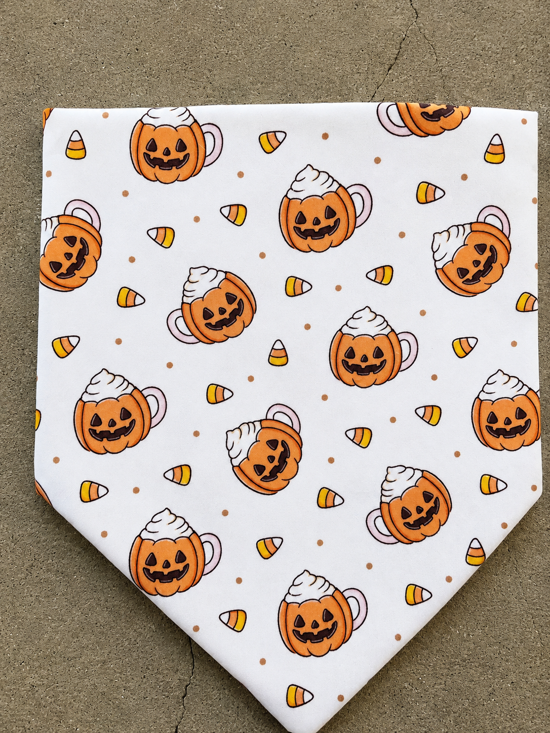 A white pet bandana featuring a Halloween pattern of jack-o'-lanterns in teacups with whipped cream and candy corn, laid flat on a gray concrete surface.